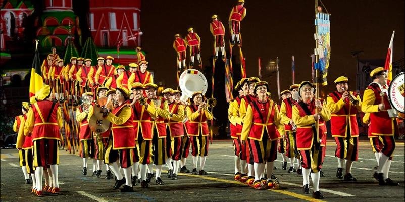 Steltenlopers en muzikanten in rood-geel kostuum op een groot plein bij avond