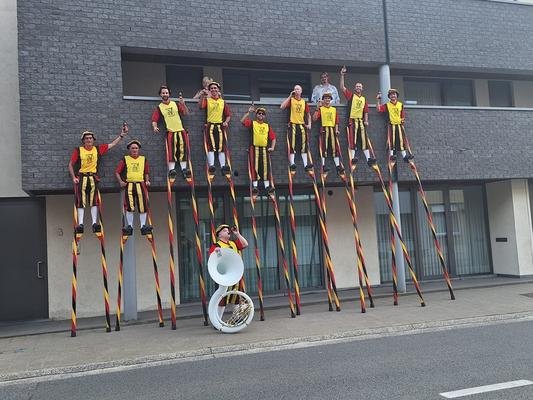 Steltenlopers zwaaien op een rij tegen een gevel, met een sousafoon ervoor