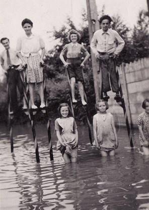 Oude zwart-witfoto van volwassenen op stelten en kinderen in het water